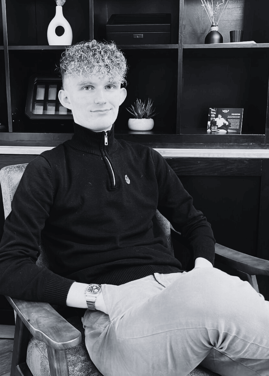Person sitting in a chair wearing a black sweater and light pants in a room with shelves and decor.