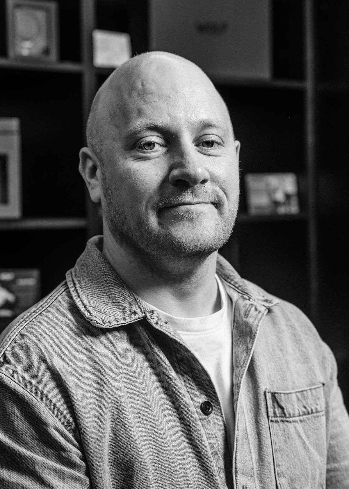 Black and white portrait of a man with a beard, wearing a denim jacket, in front of a bookshelf.